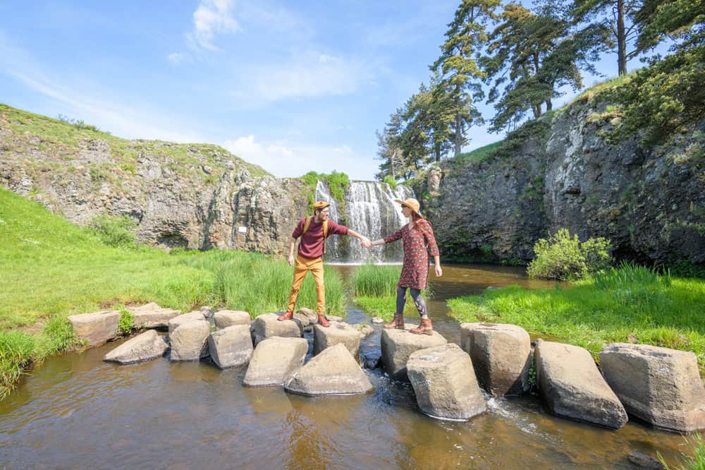 Deux personnes traversent un ruisseau avec une cascade en arrière-plan.