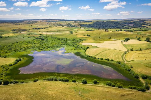 Paysage naturel avec étang, champs et collines sous un ciel bleu.