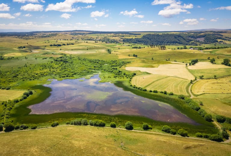 Paysage naturel avec étang, champs et collines sous un ciel bleu.